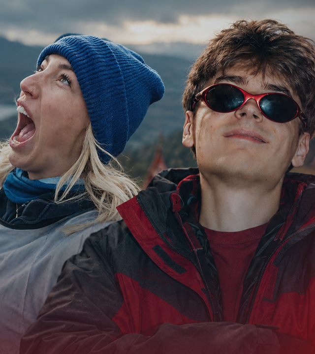 Starletnova steht links von Wichtiger in einer Berglandschaft. Sie trägt eine blaue Strickmütze, hat einen Arm ausgestreckt und blickt mit weit geöffnetem Mund nach oben. Wichtiger trägt eine rot-schwarze Jacke und eine Sonnenbrille.