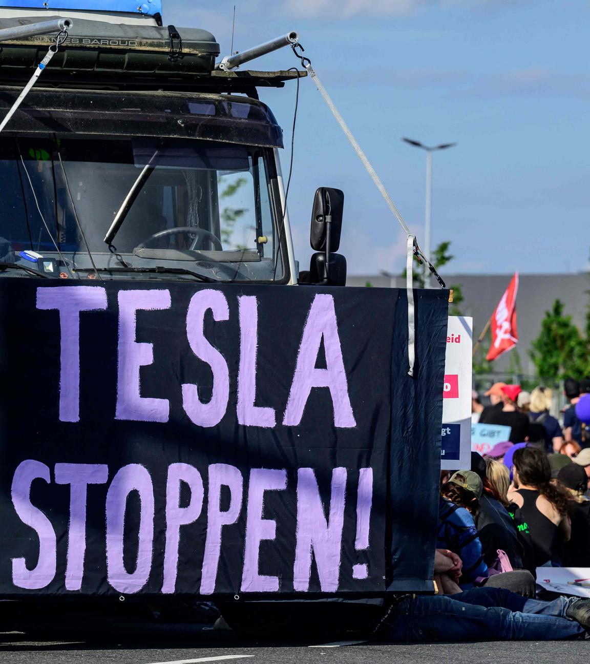Klimaaktivisten sitzen auf dem Boden und blockieren das Tesla Gelände in Grünheide. Links ist ein Banner der Aufschrift "TESLA STOPPEN" vor einen LKW gespannt.