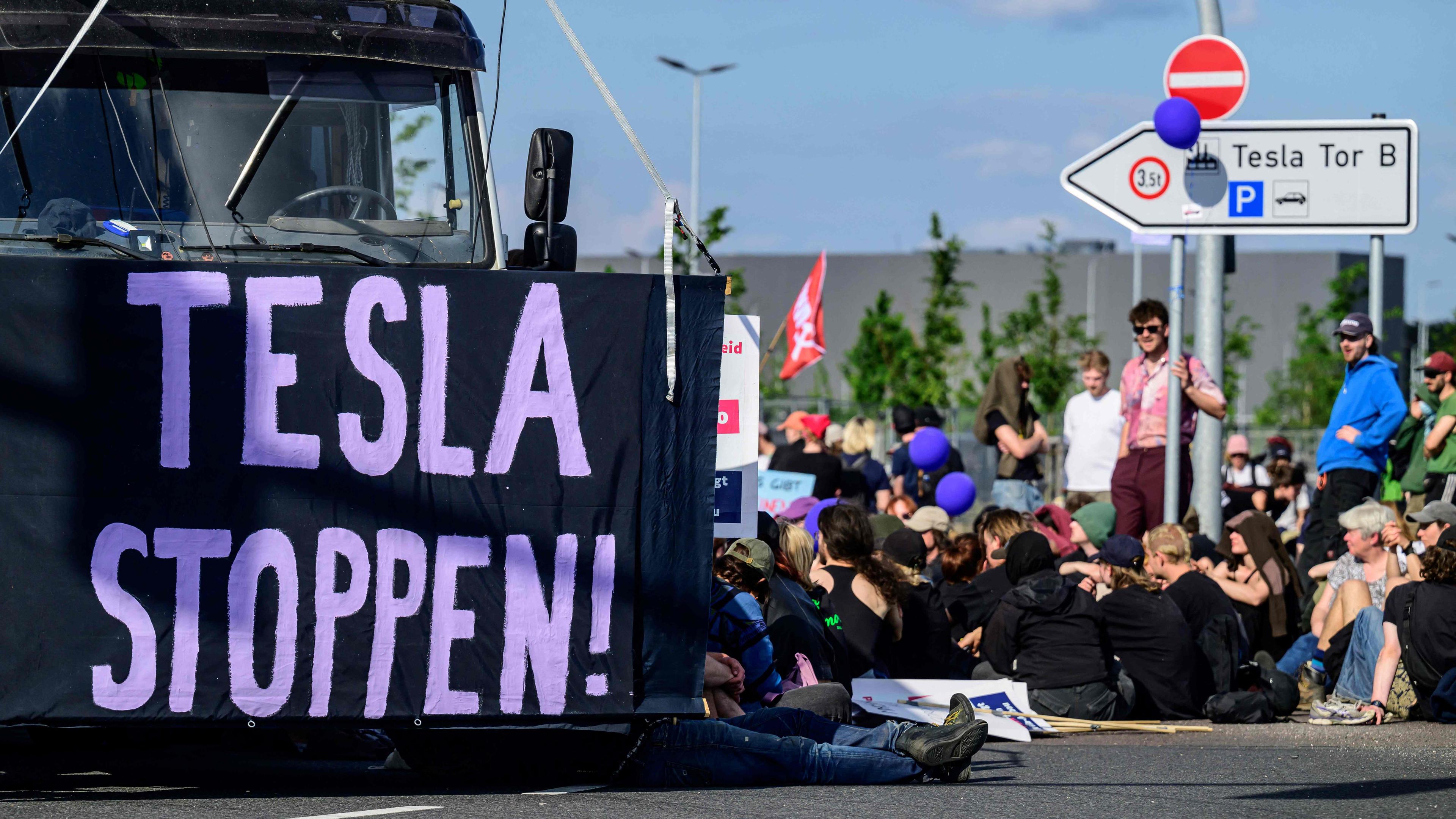 Klimaaktivisten sitzen auf dem Boden und blockieren das Tesla Gelände in Grünheide. Links ist ein Banner der Aufschrift "TESLA STOPPEN" vor einen LKW gespannt.