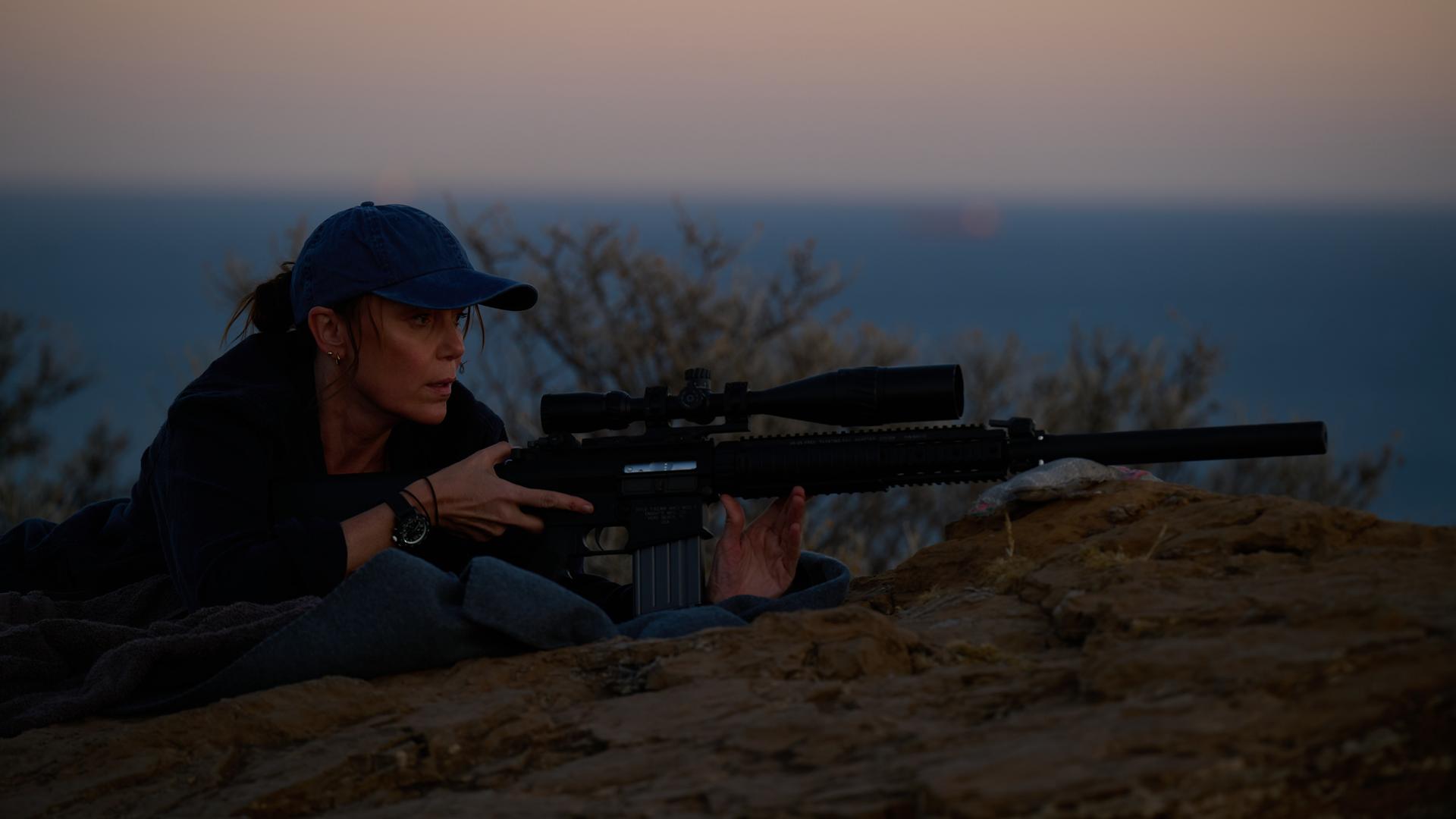 "The Assassin - Eine explosive Hochzeit": In der Dämmerung liegt Julie (Keeley Hawes) auf einem Felsen. Sie trägt eine Baseballkappe und hält ein Gewehr mit Zielfernrohr und Schalldämpfer im Anschlag.