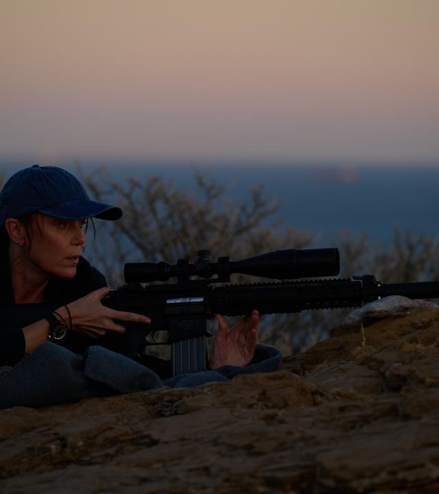 "The Assassin - Eine explosive Hochzeit": In der Dämmerung liegt Julie (Keeley Hawes) auf einem Felsen. Sie trägt eine Baseballkappe und hält ein Gewehr mit Zielfernrohr und Schalldämpfer im Anschlag.