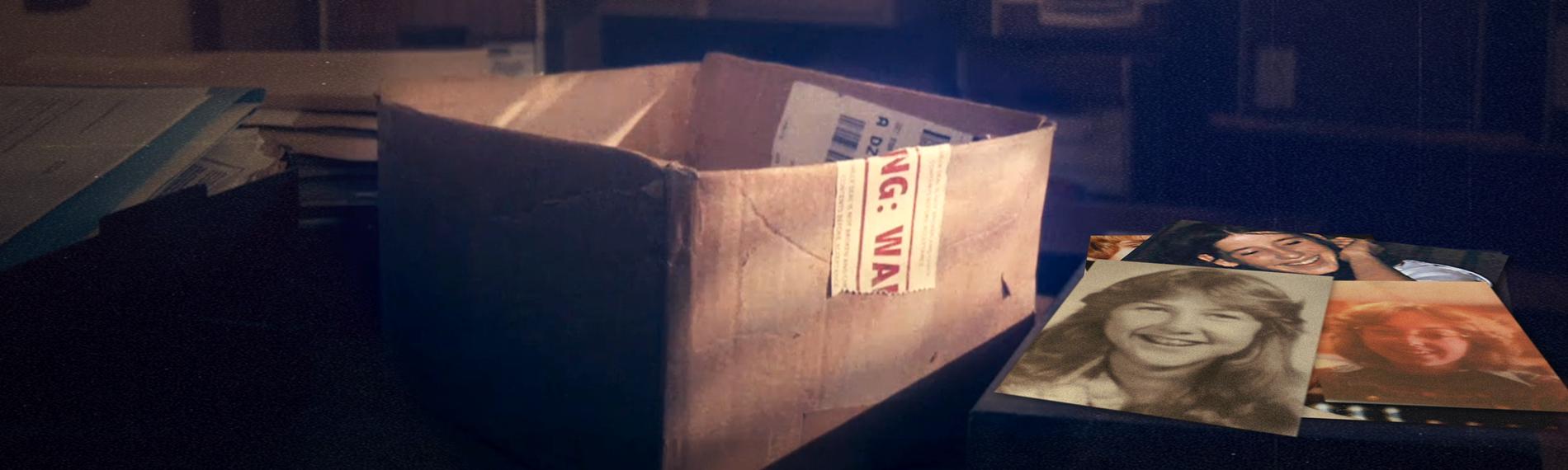 Eine offene Pappbox, im Hintergrund viele weitere Boxen, schummeriges Licht. Neben der Box liegen mehrere Fotos junger Frauen.