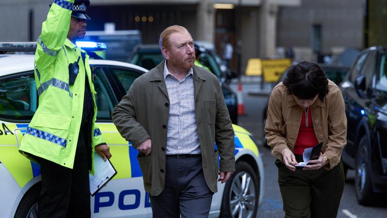 "The Chelsea Detective - Zum Wohle aller": Max (Adrian Scarborough) und Layla (Vanessa Emme) ducken unter einem Polizeiabsperrband, das ein Streifenpolizist für sie anhebt. 