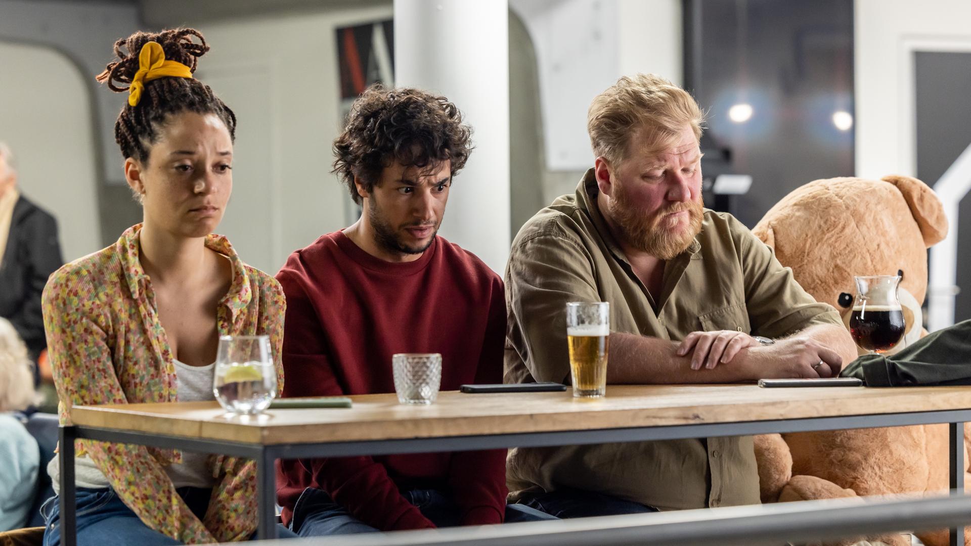 "The Club - Der Go-to-Hell-Club": Ellen (Jade Olieberg), Ziggy (Achraf Koutet) und Bert (Dominique Van Malder) sitzen betrübt an einem Tisch mit Getränken und blicken gedankenverloren vor sich hin.