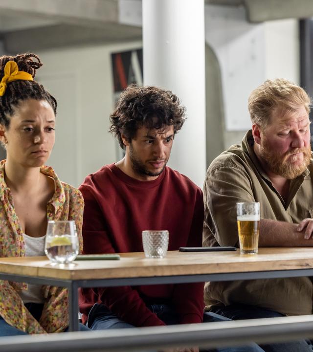 "The Club - Der Go-to-Hell-Club": Ellen (Jade Olieberg), Ziggy (Achraf Koutet) und Bert (Dominique Van Malder) sitzen betrübt an einem Tisch mit Getränken und blicken gedankenverloren vor sich hin.
