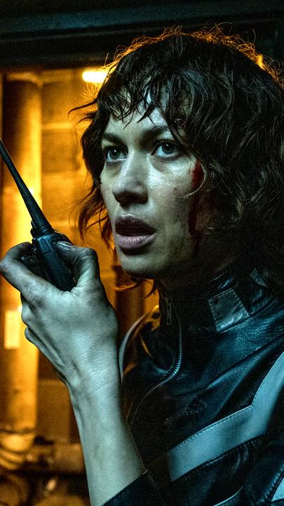 Der Kurier (Olga Kurylenko) steht in einem dunklen Keller und spricht in ein Walkie-Talkie.