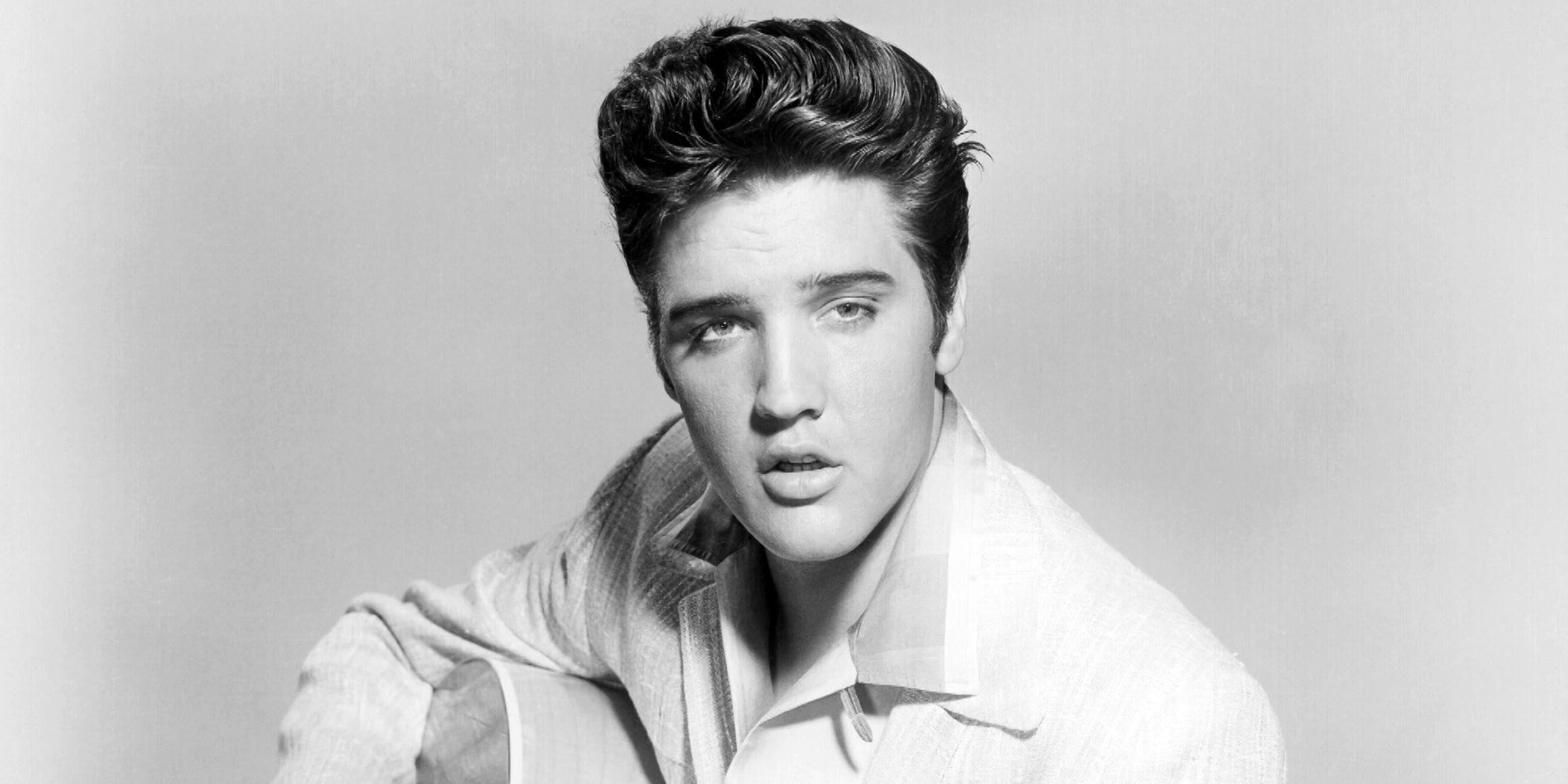  Schwarz-Weiß-Foto des jungen Elvis Presley mit Tolle und weißem Sakko. Er hält eine Akustikgitarre im Arm.