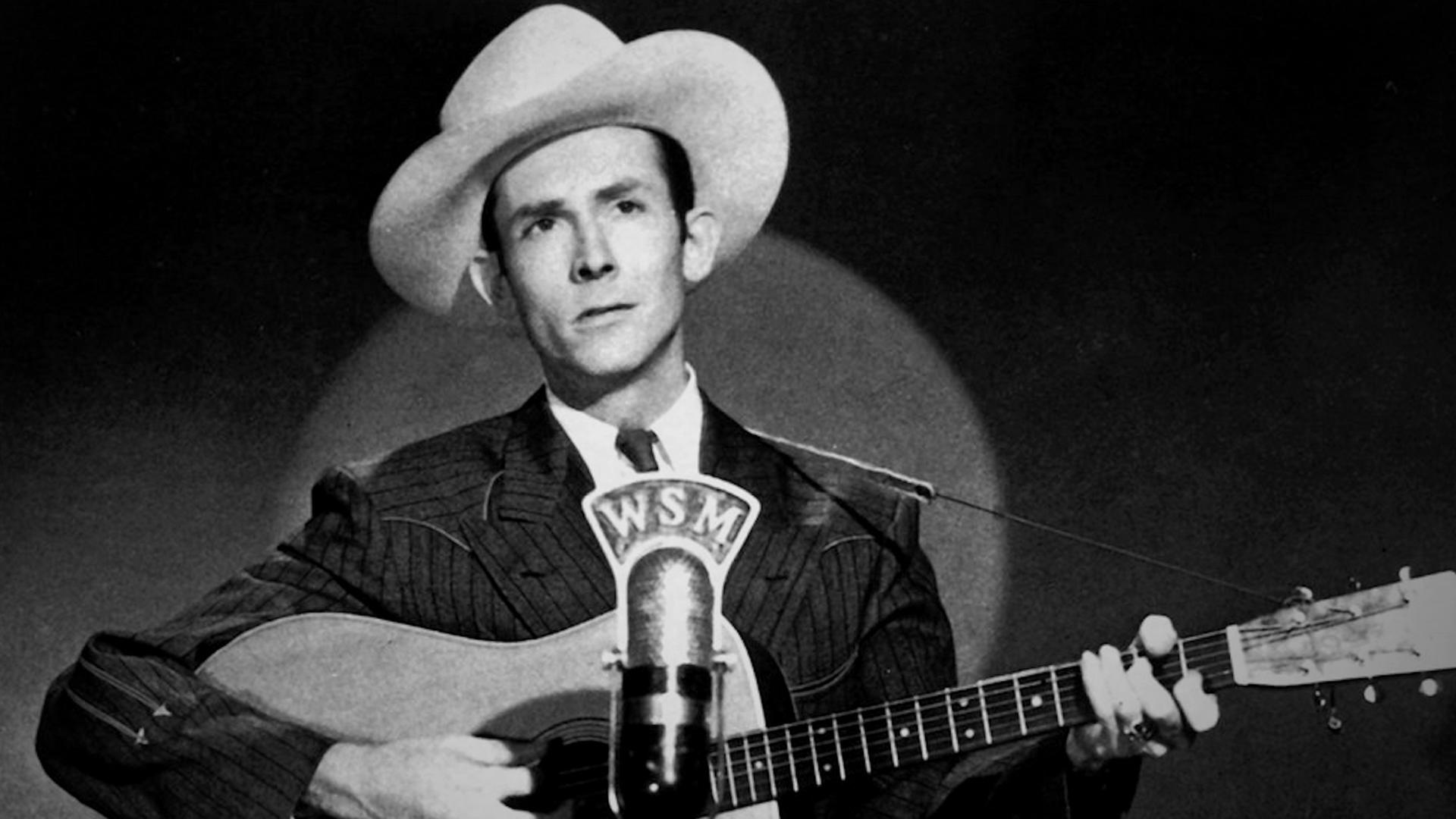  Schwarz-Weiß-Foto von Hank Williams mit Cowboyhut und Gitarre am Mikrofon.
