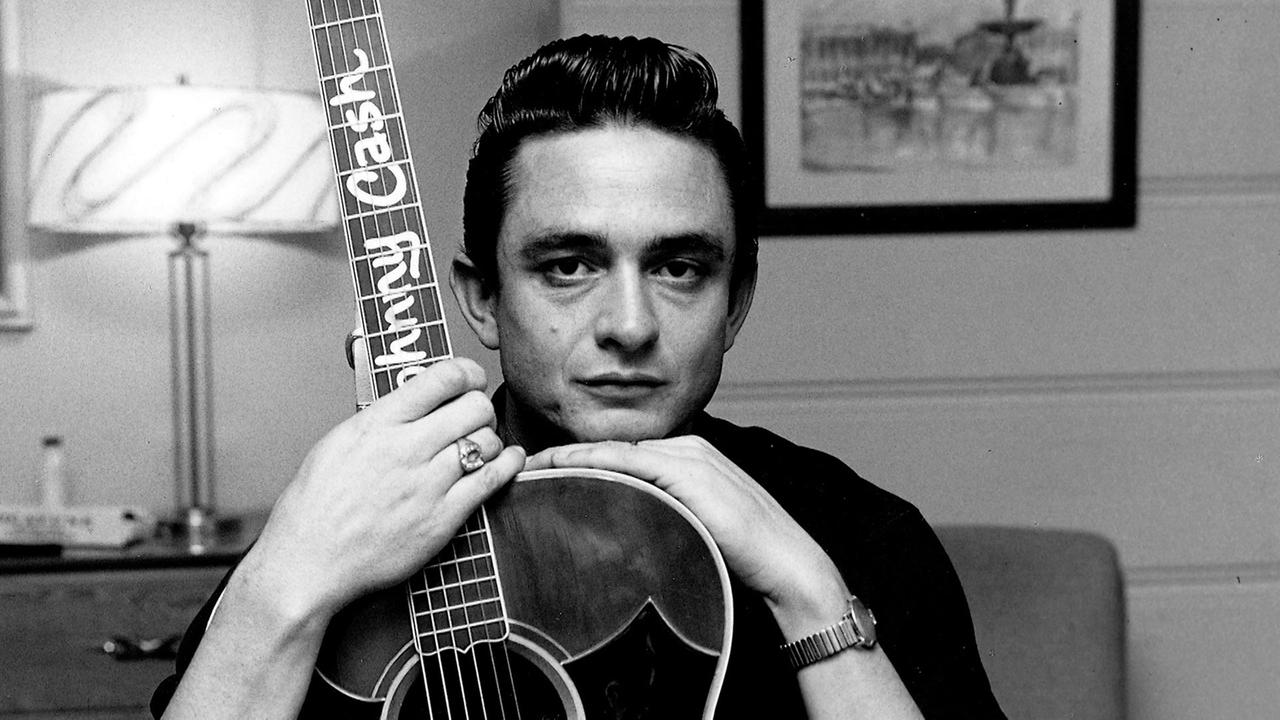  Schwarz-Weiß-Foto von Johnny Cash. Er hält eine Gitarre mit seinem Namen auf dem Gitarrenhals in den Händen. Im Hintergrund eine Lampe und ein gerahmtes Bild.