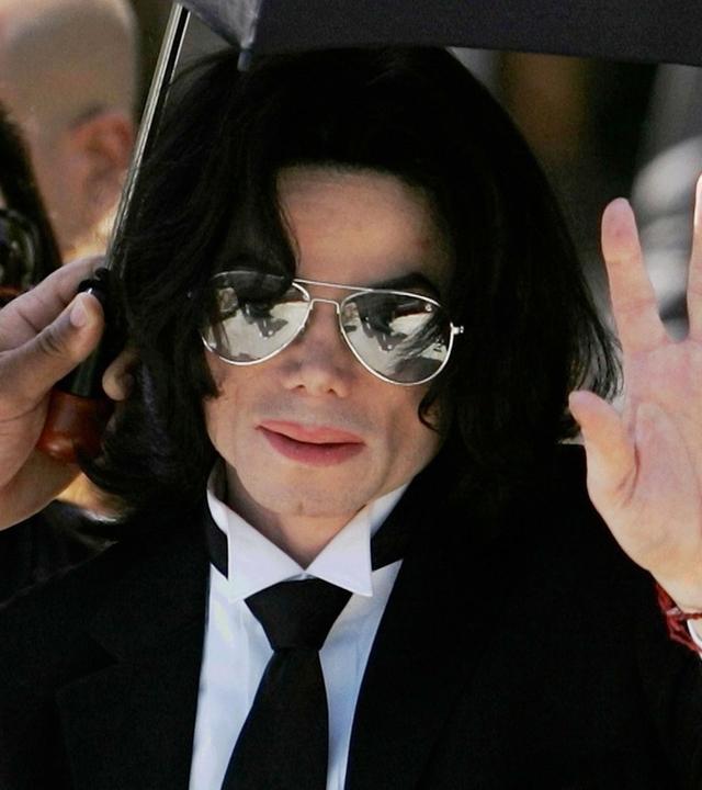 Michael Jackson mit Anzug und Krawatte inmitten einer Menschengruppe. Eine Hand hält ihm von links einen Sonnenschirm über den Kopf. Jackson hat die linke Hand erhoben.