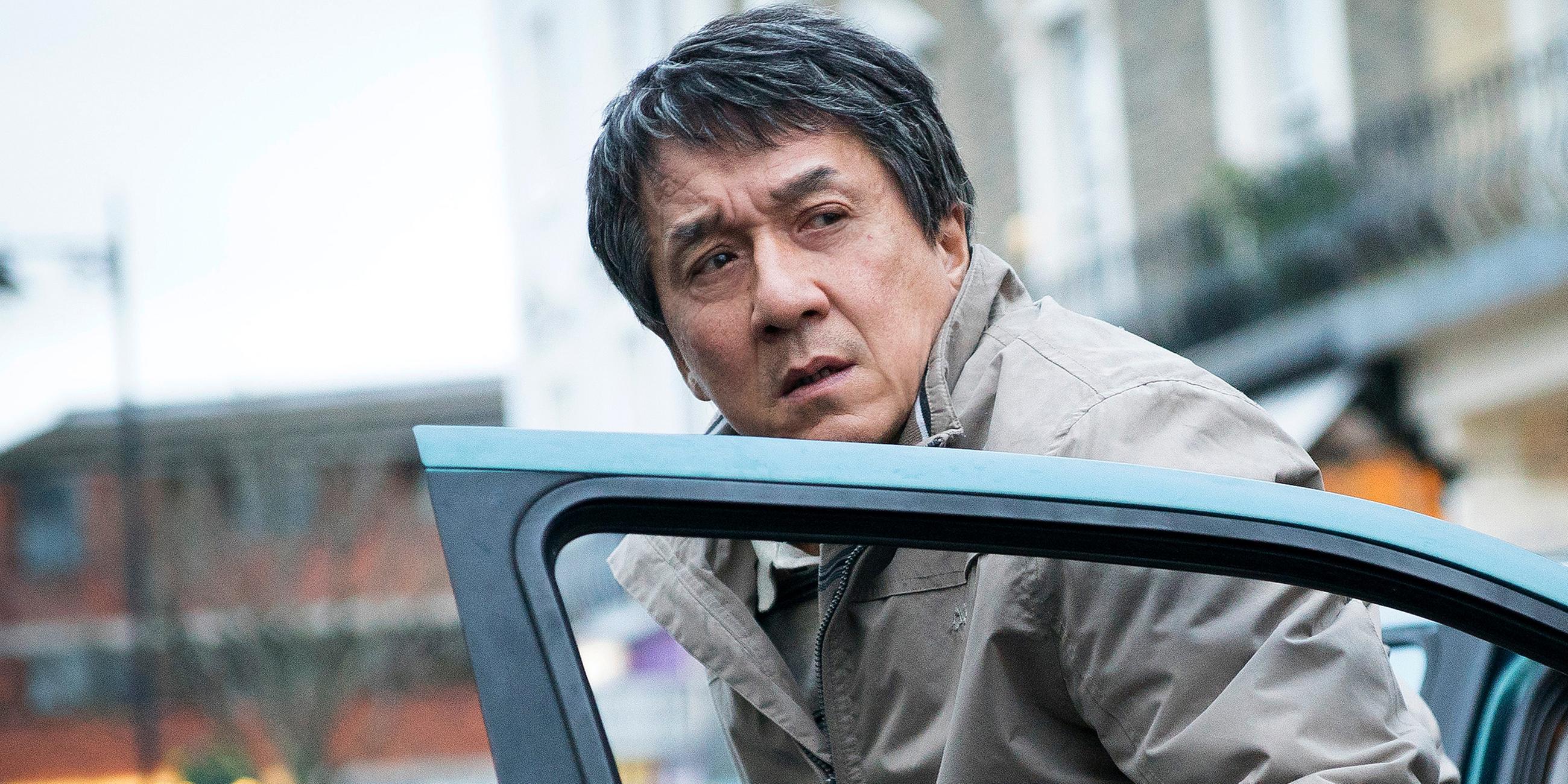 Quan Ngoc Minh (Jackie Chan) trägt eine Windjacke, ist mit ärgerlichem Blick aus dem Auto ausgestiegen und steht noch hinter der geöffneten Fahrertür. Im Hintergrund sind unscharf eine Reihe am Straßenrand geparkter Autos sowie eine Häuserfront zu erkennen.