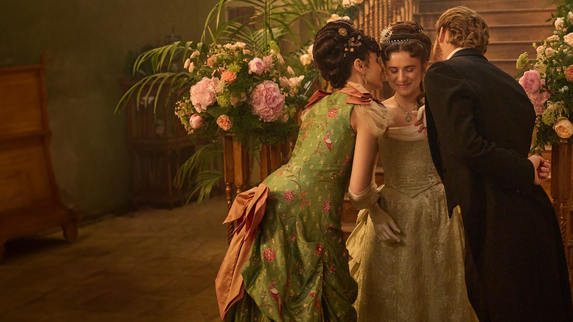 "The Forsytes - Familie verpflichtet: Der Geburtstagsball": June (Justine Emma Moore) steht zwischen ihrer Mutter Frances (Tuppence Middleton) und ihren Stiefvater Jolyon Jr. (Danny Griffin) in einer Empfangshalle vor einer großen hölzernen Treppe, die mit opulenten Blumenschmuck verziert ist.