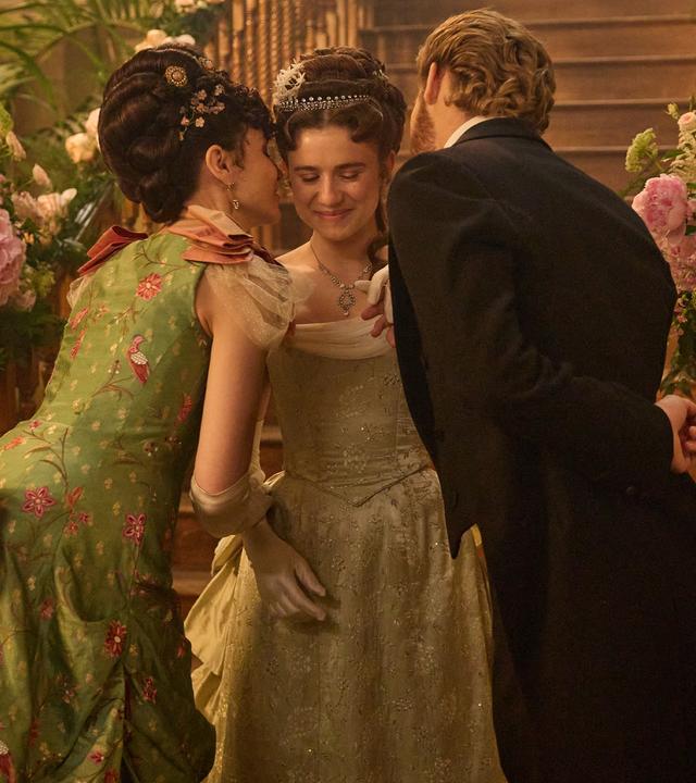 "The Forsytes - Familie verpflichtet: Der Geburtstagsball": June (Justine Emma Moore) steht zwischen ihrer Mutter Frances (Tuppence Middleton) und ihren Stiefvater Jolyon Jr. (Danny Griffin) in einer Empfangshalle vor einer großen hölzernen Treppe, die mit opulenten Blumenschmuck verziert ist.