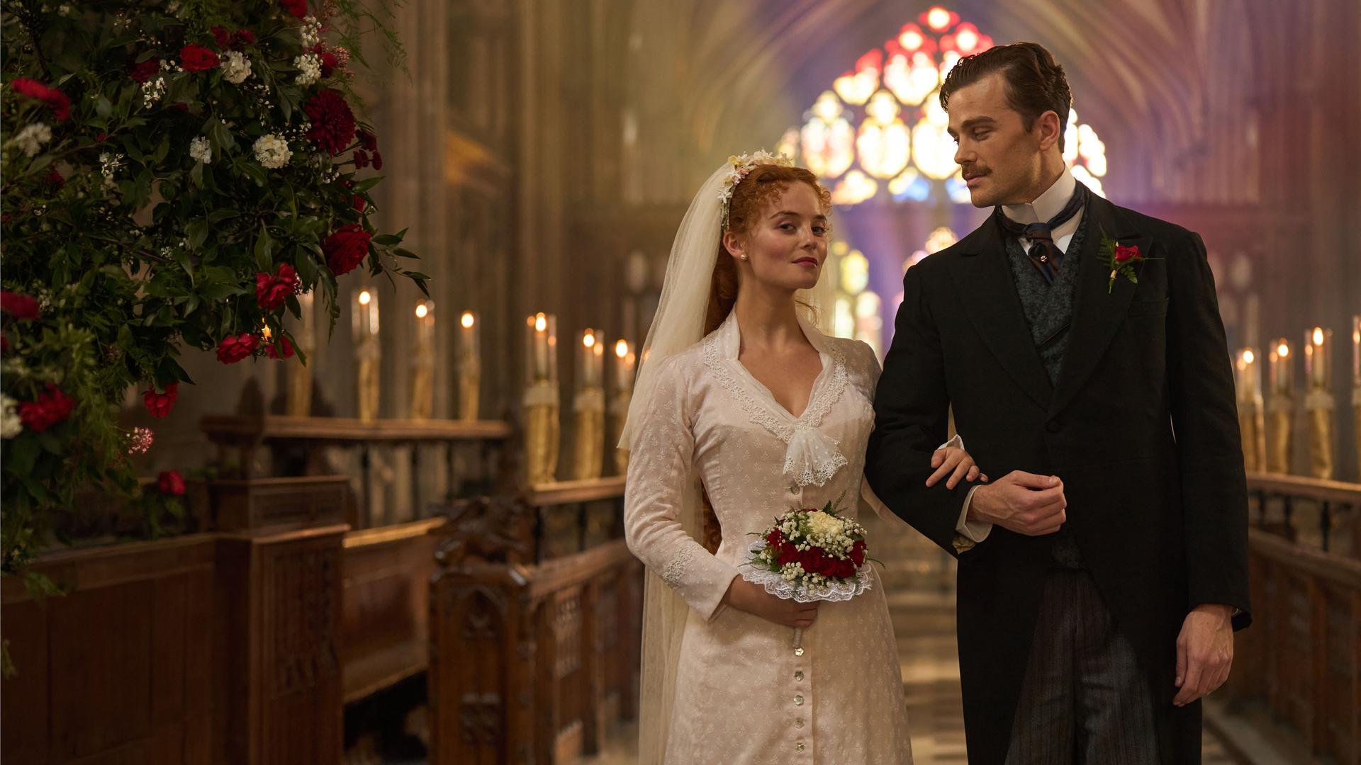"The Forsytes - Familie verpflichtet: Loyalität oder Liebe": Irene (Millie Gibson) und Soames (Joshua Orpin) stehen nebeneinander in einer Kirche. Im Hintergrund leuchten in einer Reihe aufgestellte Kerzen und, farbiges Sonnlicht fällt durch die bunten Kirchenfenster. Sie trägt ein schlichtes weißes Kleid mit einem weißen langen Schleier im Haar und hält einem Blumenstrauß in der Hand. Soames trägt einen schwarzen Anzug mit Nadelstreifenhose und dazu passender Weste. Irene hat sich bei Soames eingehackt und blickt nach vorne, während er sie ansieht.