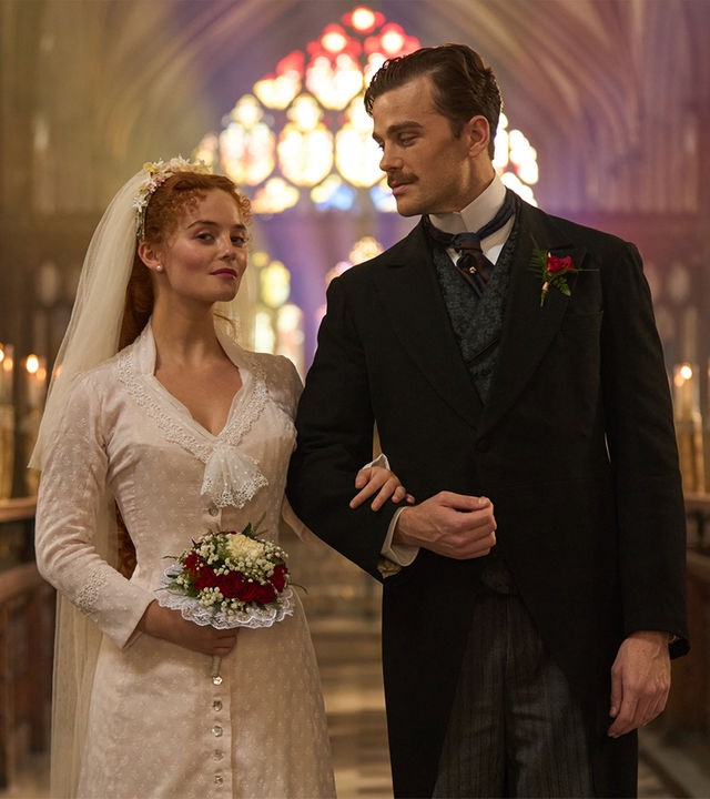 "The Forsytes - Familie verpflichtet: Loyalität oder Liebe": Irene (Millie Gibson) und Soames (Joshua Orpin) stehen nebeneinander in einer Kirche. Im Hintergrund leuchten in einer Reihe aufgestellte Kerzen und, farbiges Sonnlicht fällt durch die bunten Kirchenfenster. Sie trägt ein schlichtes weißes Kleid mit einem weißen langen Schleier im Haar und hält einem Blumenstrauß in der Hand. Soames trägt einen schwarzen Anzug mit Nadelstreifenhose und dazu passender Weste. Irene hat sich bei Soames eingehackt und blickt nach vorne, während er sie ansieht.