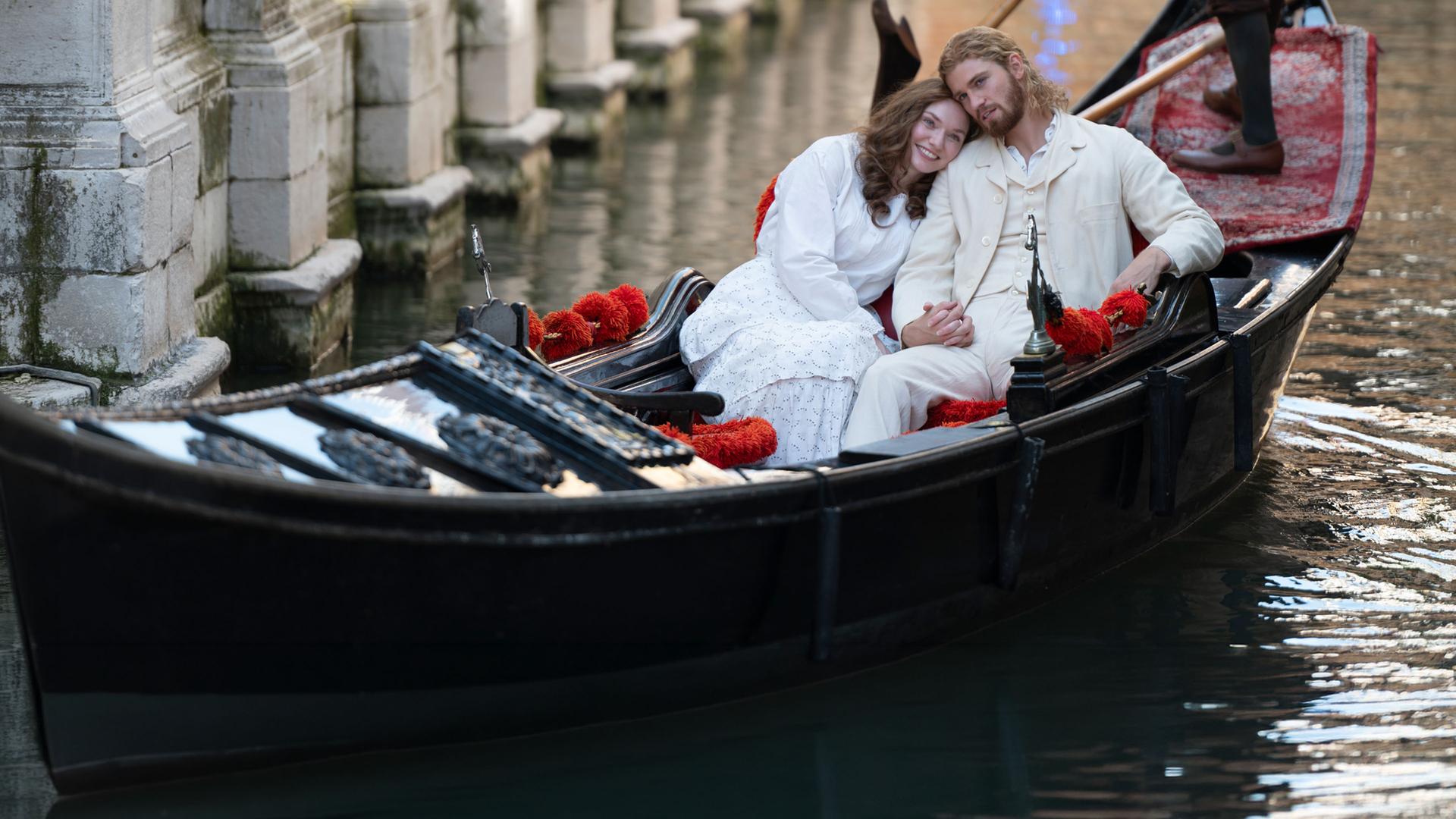 "The Forsytes - Familie verpflichtet: Heimliche Begierden": In einem Kanal in Venedig in einer Gondel: Luisa (Eleanor Tomlinson) lehnt ihren Kopf an Joylons (Danny Griffin) Schulter, beide sind weiß gekleidet. Hinter ihnen steht der Gondolière, der bis zur Hüfte zu erkennen ist.