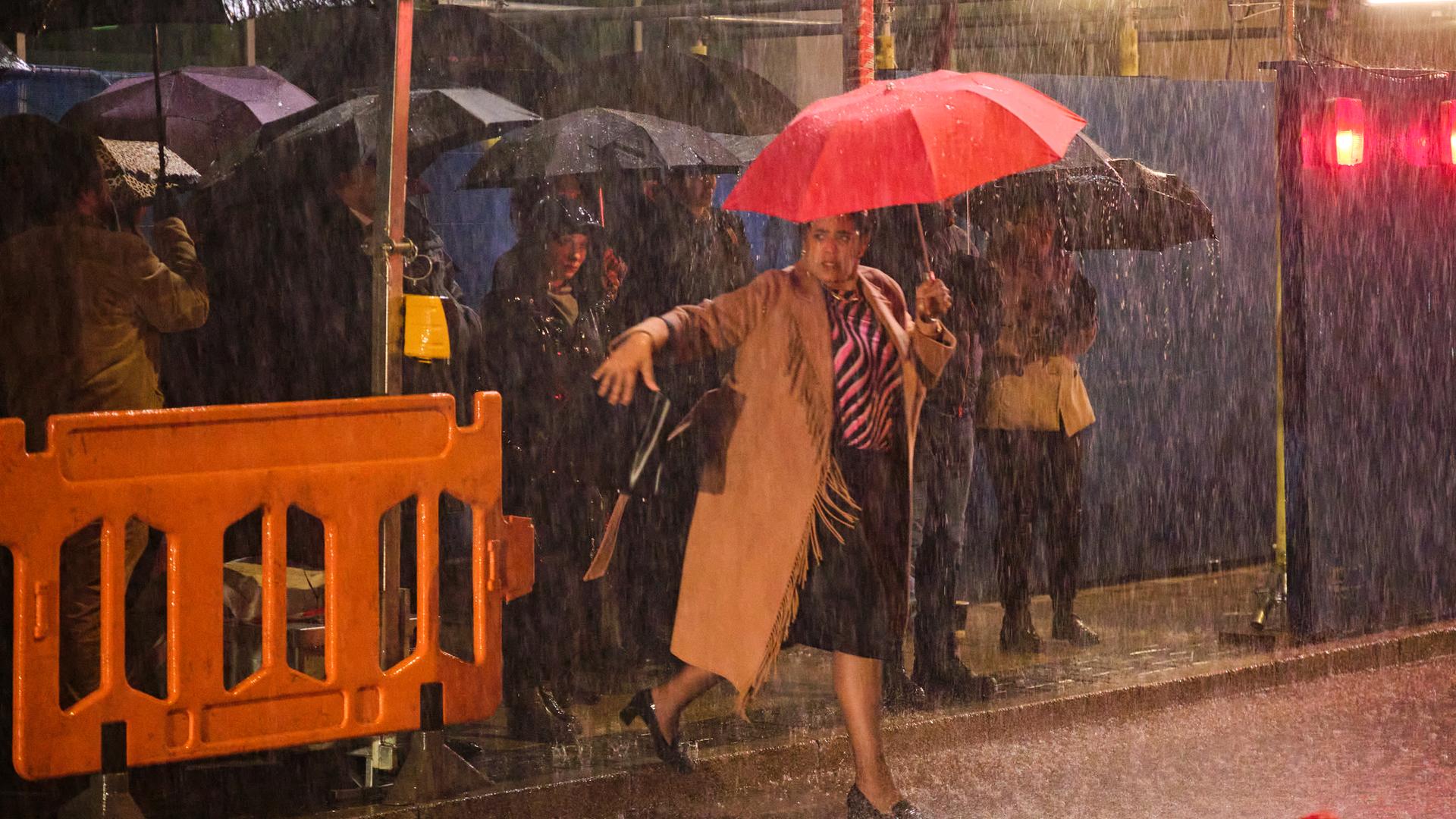 Anwältin Belinda Grey, die einen roten Schirm über sich hält, läuft nachts bei Regen vor einer Baustelle auf die Fahrbahn der Straße. Im Hintergrund befinden sich weitere Personen (Komparsen) mit schwarzen Schirmen auf dem Gehweg.