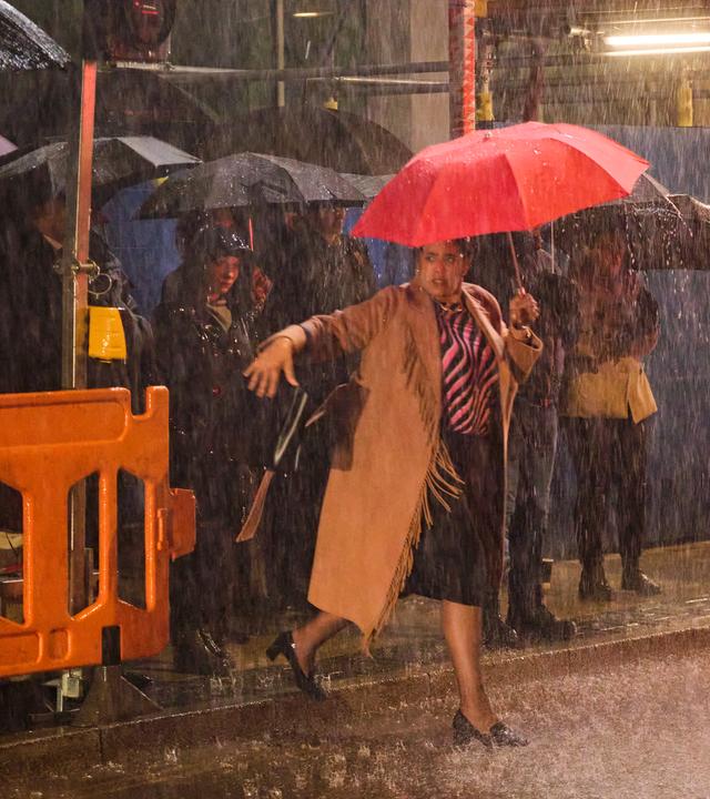 Anwältin Belinda Grey, die einen roten Schirm über sich hält, läuft nachts bei Regen vor einer Baustelle auf die Fahrbahn der Straße. Im Hintergrund befinden sich weitere Personen (Komparsen) mit schwarzen Schirmen auf dem Gehweg.