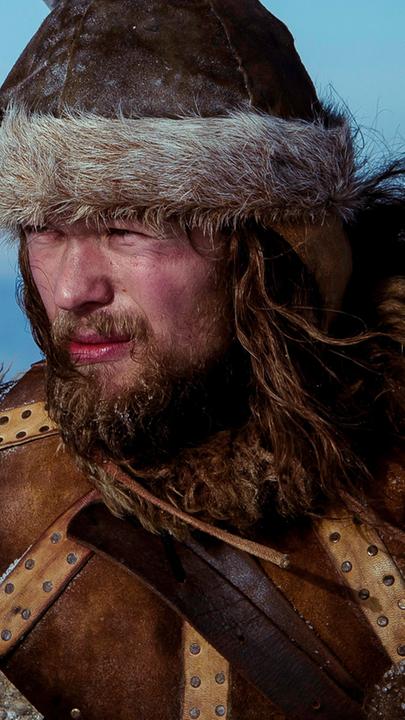 Skjervald (Jakob Oftebro) hält einen Speer in der Hand und sieht nach vorne. Auf dem Rücken trägt er ein Baby.