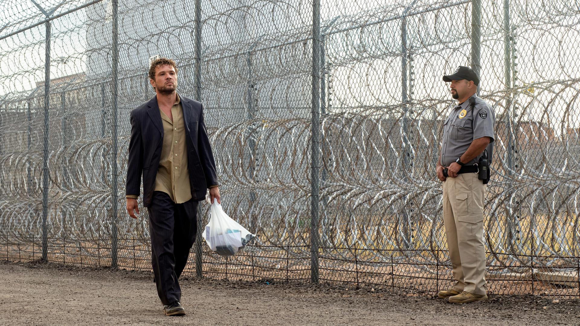 The Locksmith  Der Spezialist: Miller (Ryan Phillippe) läuft mit einer gefüllten Plastiktüte und ernstem Blick an einem hohen Maschendrahtzaun mit Stacheldraht entlang, während ein uniformierter Polizist in der Nähe steht.