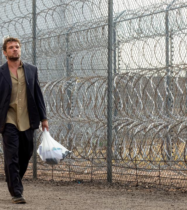 The Locksmith  Der Spezialist: Miller (Ryan Phillippe) läuft mit einer gefüllten Plastiktüte und ernstem Blick an einem hohen Maschendrahtzaun mit Stacheldraht entlang, während ein uniformierter Polizist in der Nähe steht.