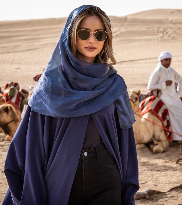 Eine Frau in dunklem Umhang und Sonnenbrille steht in einer Wüste vor einer Reihe sitzender Dromedare. Im Hintergrund sitzt ein Mann in weißer Kleidung auf einem Dromedar.