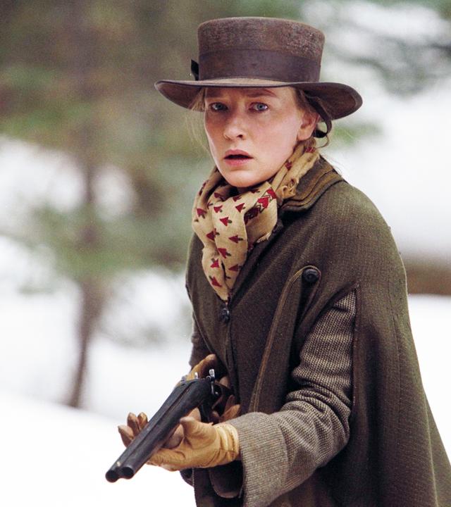 "The Missing": Mit angespanntem Gesichtsausdruck hält Maggie Gilkeson (Cate Blanchett) eine Schrotflinte im Anschlag. Sie ist bekleidet mit einem warmen Übermantel sowie mit Handschuhen, Schaltuch und Hut. Im Hintergrund sind unscharf vereinzelte Bäumen und Schnee zu erkennen.
