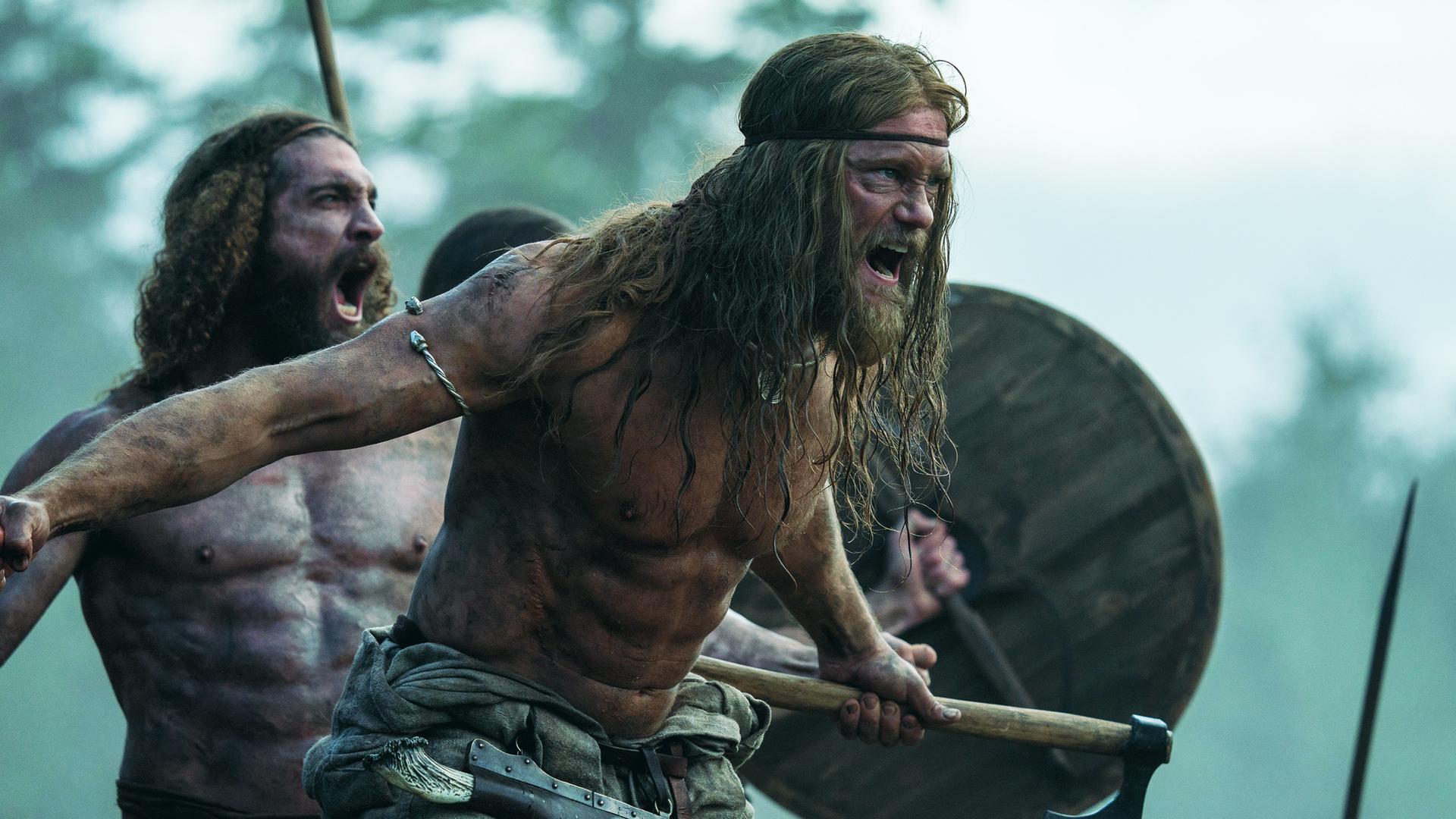 Halbnahe: Amleth (Alexander Skarsgård), muskulös und oberkörperfrei, steht schreiend mit gezücktem Beil vor einem weiteren brüllenden Mann (Komparse) von ähnlicher Statur. Dahinter ist durch ein erhobenes Holzschild und eine Lanze eine weitere Person zu erahnen. Im Hintergrund sind unscharf Bäume zu erkennen.