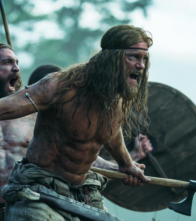 Halbnahe: Amleth (Alexander Skarsgård), muskulös und oberkörperfrei, steht schreiend mit gezücktem Beil vor einem weiteren brüllenden Mann (Komparse) von ähnlicher Statur. Dahinter ist durch ein erhobenes Holzschild und eine Lanze eine weitere Person zu erahnen. Im Hintergrund sind unscharf Bäume zu erkennen.
