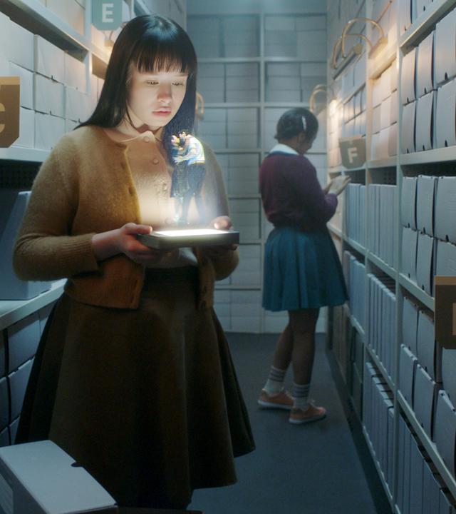 "The Ordinaries": Paula (Fine Sendel) steht im Gang eines Archivs und schaut auf ein Hologramm, welches sie in ihren Händen hält.  