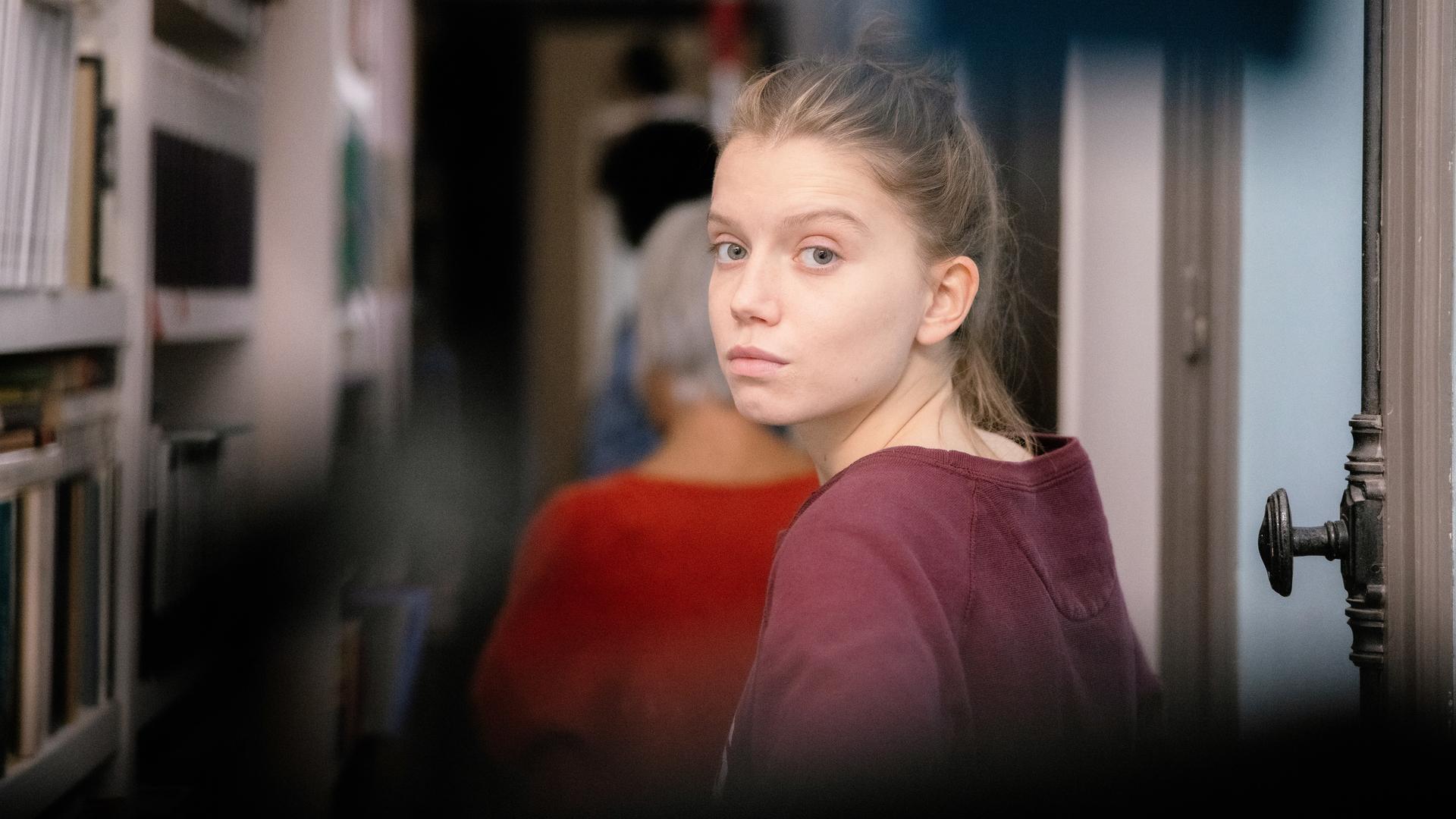 "The Perfect Mother (3)": Nahaufnahme. Anya (Eden Ducourant) läuft in einer Wohnung in einen Flur. Sie schaut sich um und blickt verzweifelt direkt in die Kamera. Vor ihr läuft, unscharf zu erkennen, ihre Großmutter Élisabeth (Frédérique Tirmont).