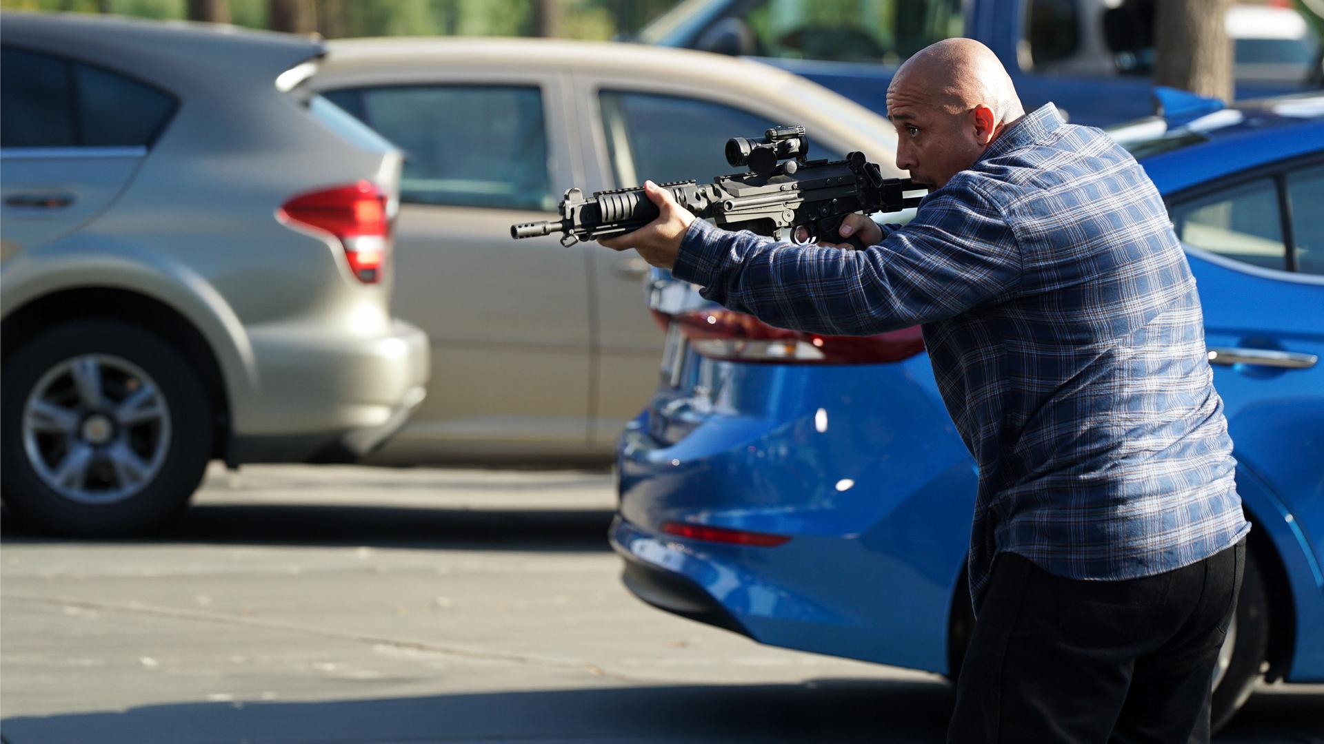 "The Rookie - Der Tausch": Außenaufnahme auf einem Parkplatz. Ein kahlköpfiger Mann hält mit beiden Händen ein Gewehr abschussbereit nach vorne.