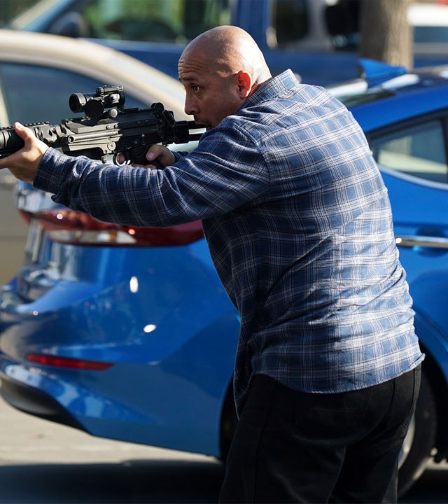 "The Rookie - Der Tausch": Außenaufnahme auf einem Parkplatz. Ein kahlköpfiger Mann hält mit beiden Händen ein Gewehr abschussbereit nach vorne.