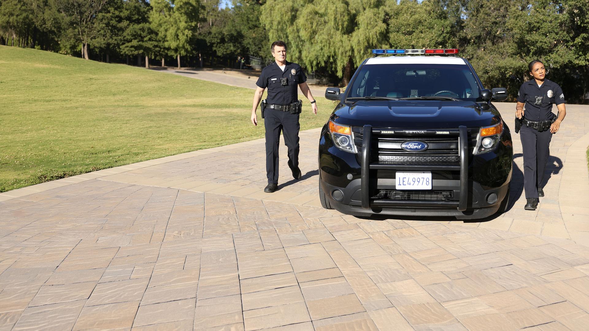 "The Rookie - Ein gefallener Stern": Nolan (Nathan Fillion) und Harper (Mekia Cox) laufen neben einem Streifenwagen her, der in einer Einfahrt geparkt ist.