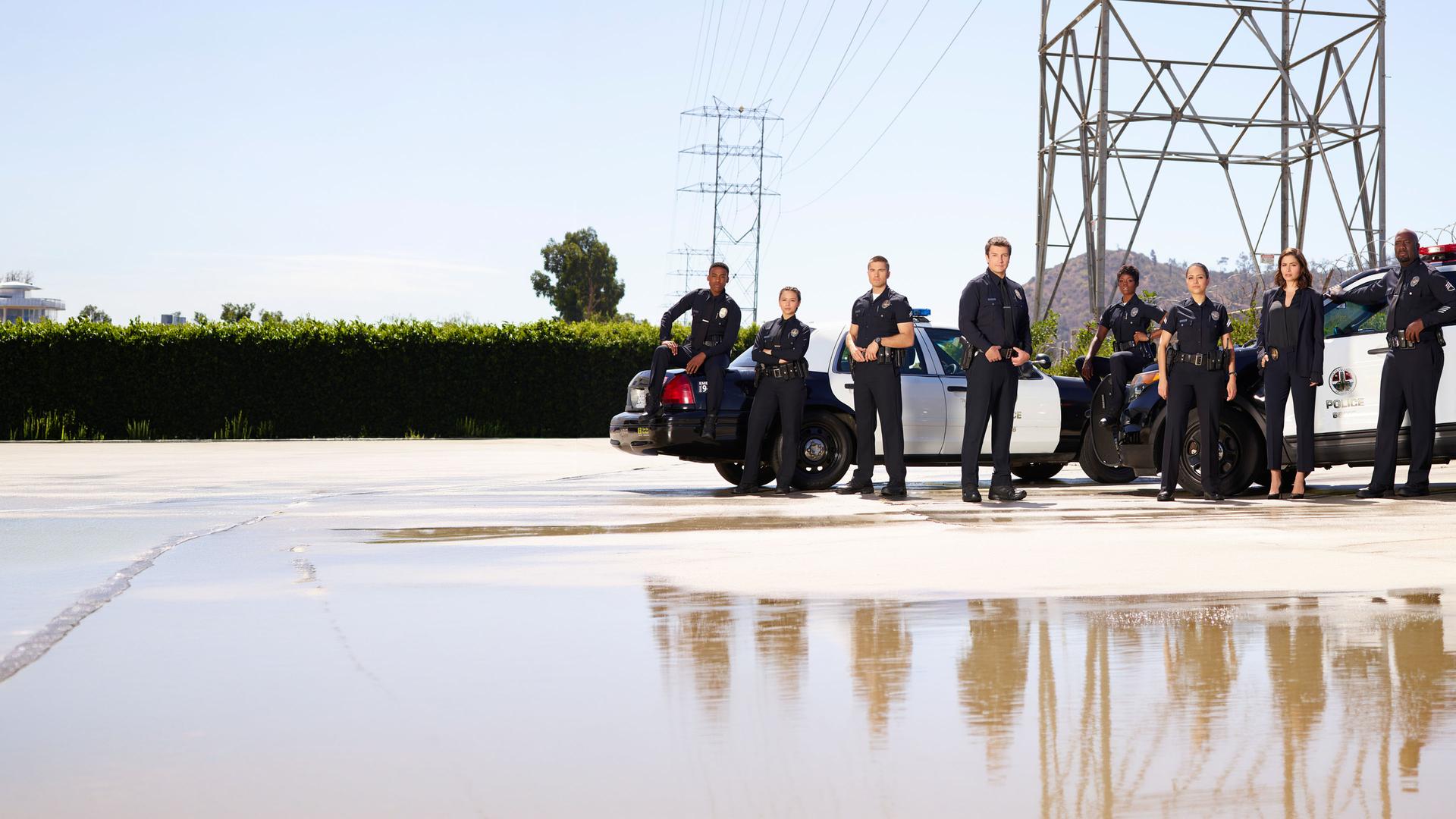 "The Rookie - Ein neuer Anfang": Sergeant Wade Grey (Richard T. Jones), Tim Bradford (Eric Winter), Talia Bishop (Afton Williamson), Angela Lopez (Alyssa Diaz), Zoe Andersen (Mercedes Mason), Lucy Chen (Melissa O'Neil), John Nolan (Nathan Fillion) und Jackson West (Titus Makin) stehen in Polizeiuniform nebeneinander. Im Hintergrund sind zwei Polizeiautos geparkt.