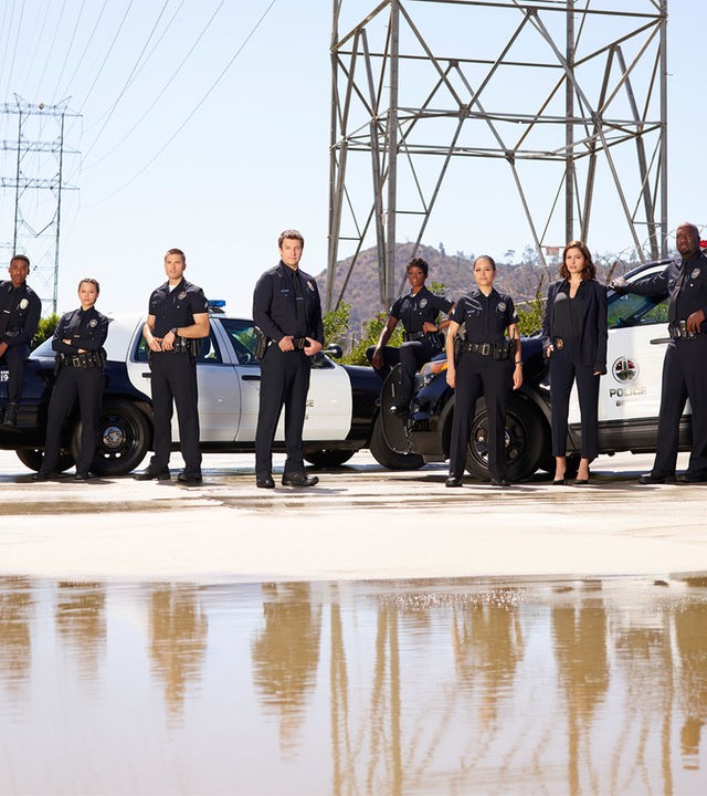 "The Rookie - Ein neuer Anfang": Sergeant Wade Grey (Richard T. Jones), Tim Bradford (Eric Winter), Talia Bishop (Afton Williamson), Angela Lopez (Alyssa Diaz), Zoe Andersen (Mercedes Mason), Lucy Chen (Melissa O'Neil), John Nolan (Nathan Fillion) und Jackson West (Titus Makin) stehen in Polizeiuniform nebeneinander. Im Hintergrund sind zwei Polizeiautos geparkt.