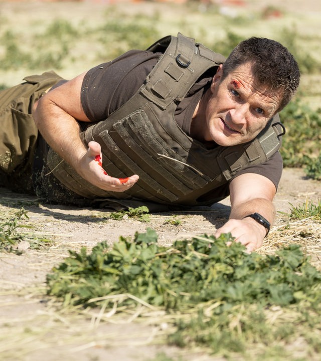 "The Rookie - Fluchtplan": Nolan (Nathan Fillion) liegt verletzt auf dem sandigen Boden und blickt angestrengt geradeaus. An seiner Hand, die er vor seinen Oberkörper hält, befindet sich Blut.