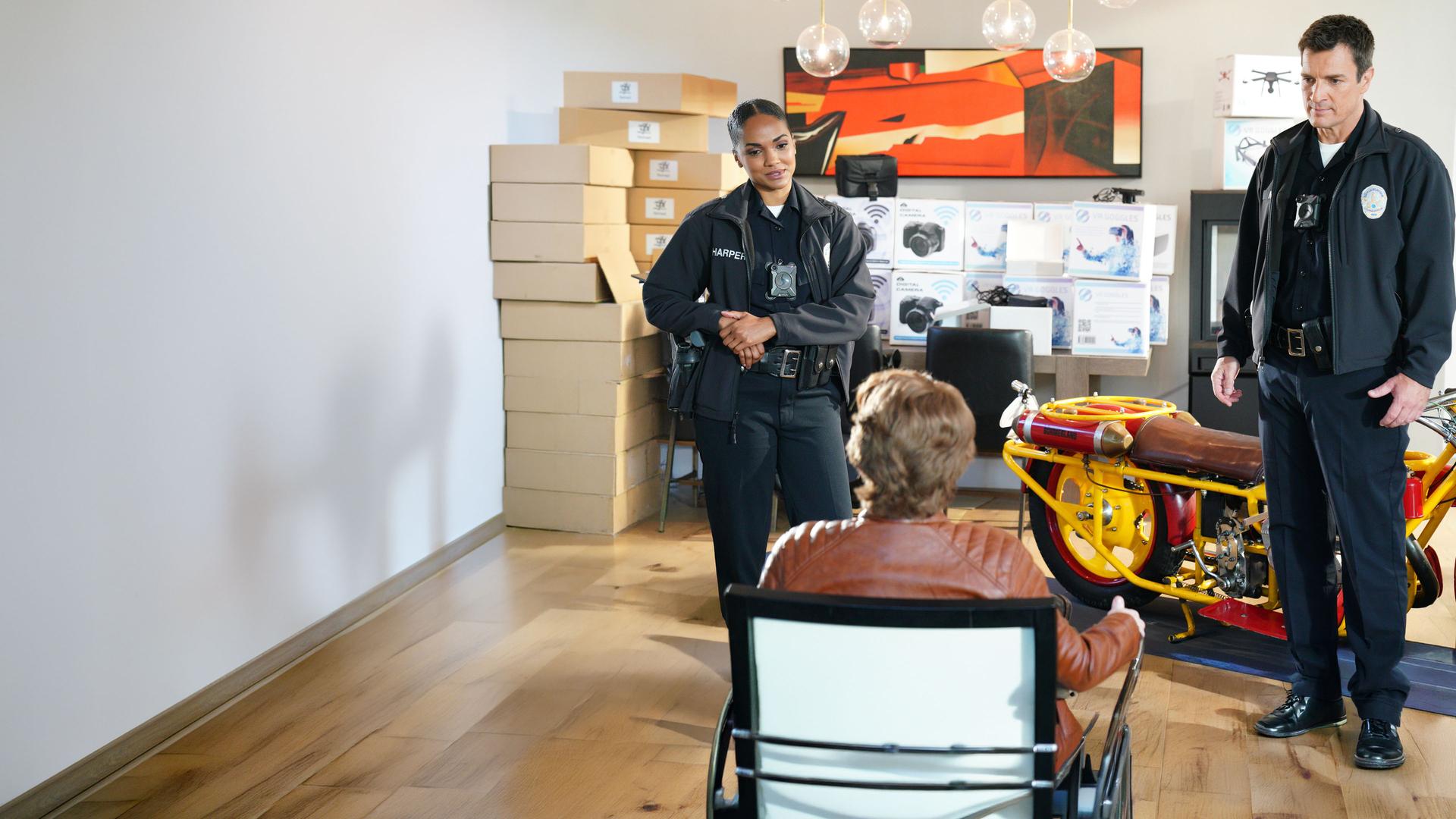 "The Rookie - Hand ab”: John Nolan (Nathan Fillion) und Nyla Harper (Mekia Cox) stellen den Identitätsdieb in seinem Wohnzimmer zwischen Kartons mit Kameras und Drohnen, sowie einem Motorrad.