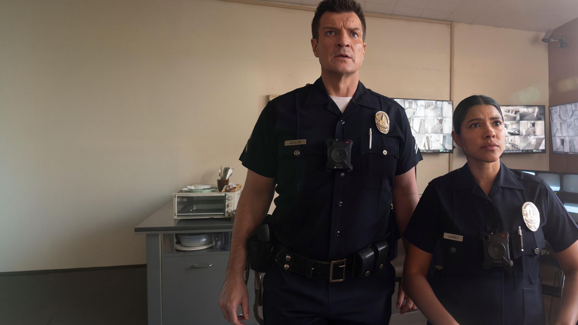 "The Rookie - Westview": Officer John Nolan (Nathan Fillion) und Celina Juarez (Lisseth Chavez) stehen nebeneinander in einem Überwachungsraum. Beide tragen dunkle Polizeiuniformen und blicken fragend nach vorn. Im Hintergrund hängen mehrere Bildschirme mit unterschiedlichen Überwachungsaufnahmen an den Wänden.