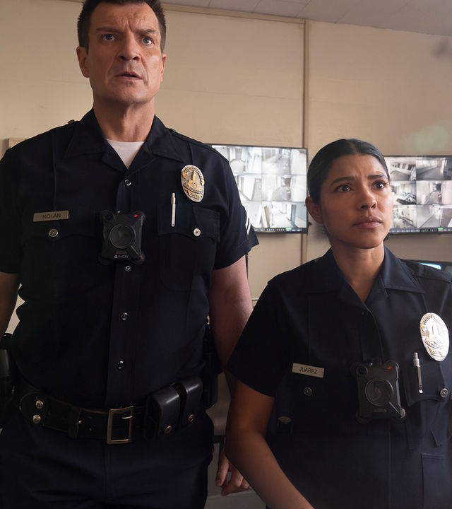 "The Rookie - Westview": Officer John Nolan (Nathan Fillion) und Celina Juarez (Lisseth Chavez) stehen nebeneinander in einem Überwachungsraum. Beide tragen dunkle Polizeiuniformen und blicken fragend nach vorn. Im Hintergrund hängen mehrere Bildschirme mit unterschiedlichen Überwachungsaufnahmen an den Wänden.
