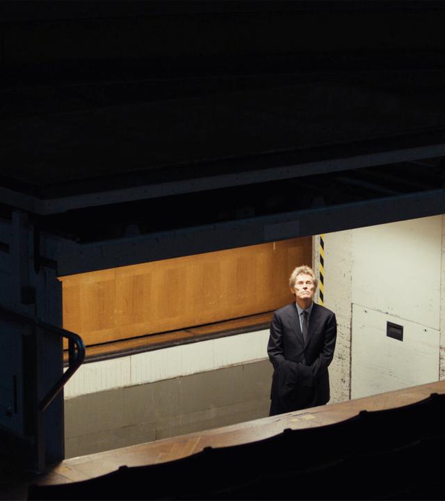 Blick von schräg oben in einen hell erleuchteten Bühnenaufzug. Darin steht ein Mann im Anzug und schaut nach oben.