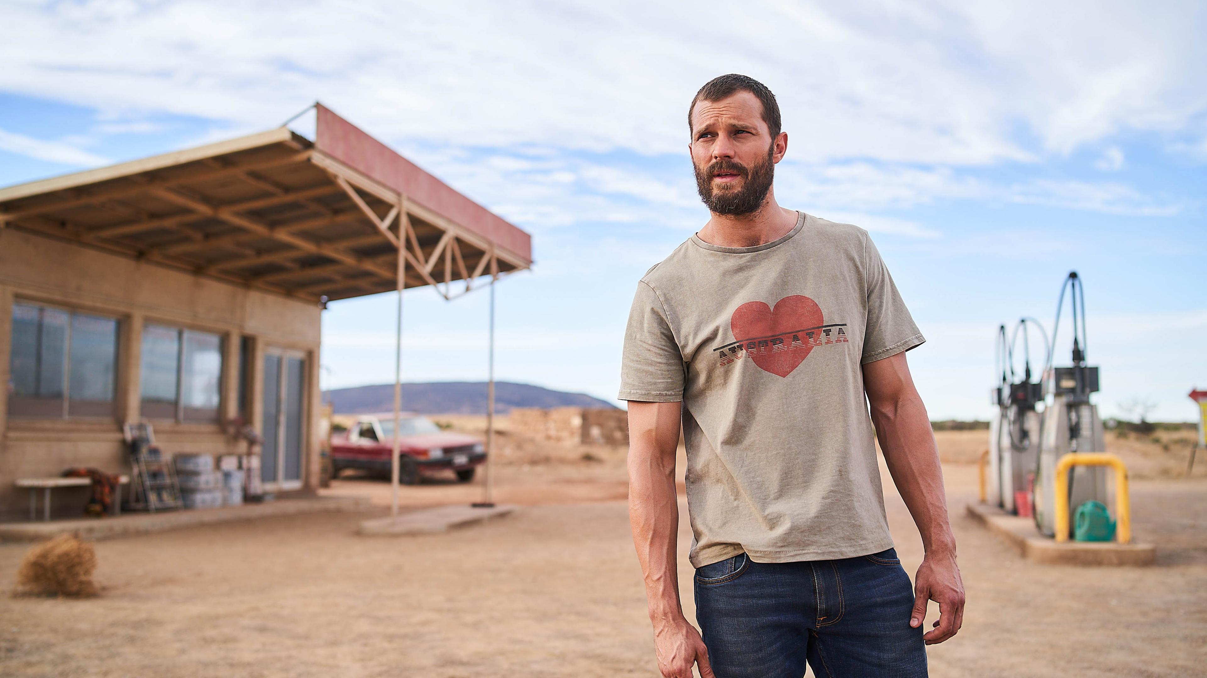 "The Tourist - Duell im Outback": Der Fremde (Jamie Dornan) steht breitbeinig und mit leicht verwirrtem Gesichtsausdruck auf dem Gelände einer kleinen Tankstelle im Outback. Er trägt eine Jeans und ein T-Shirt mit einem roten Herz und dem Schriftzug "Australia".