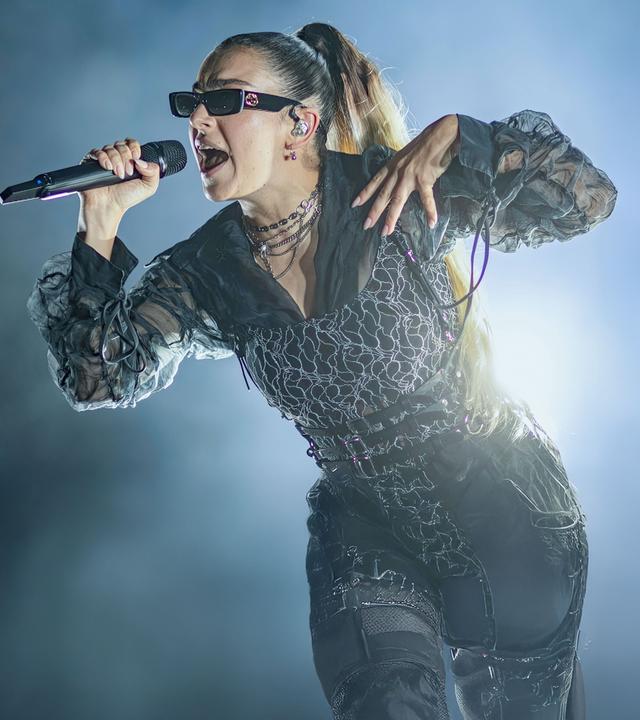Charli xcx trägt eine Sonnenbrille und singt in ein Mikrofon. Der Hintergrund ist dunkel, auf einer Seite ist Nebel.