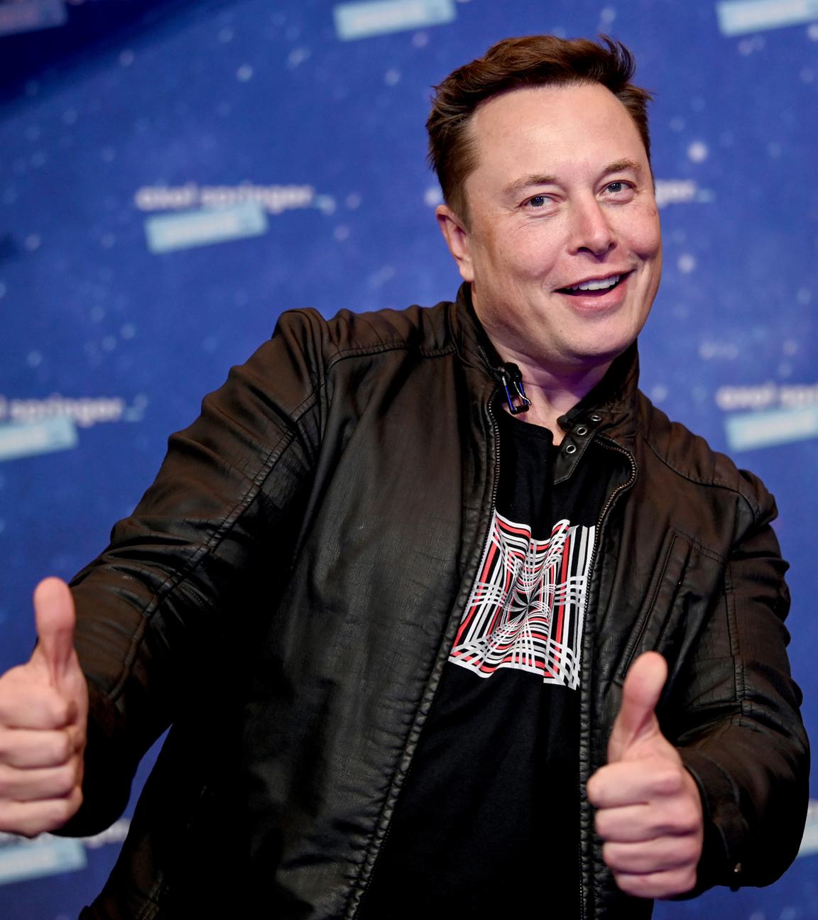 Elon Musk steht vor einer blauen Wand und zeigt zwei Daumen hoch in die Kamera.