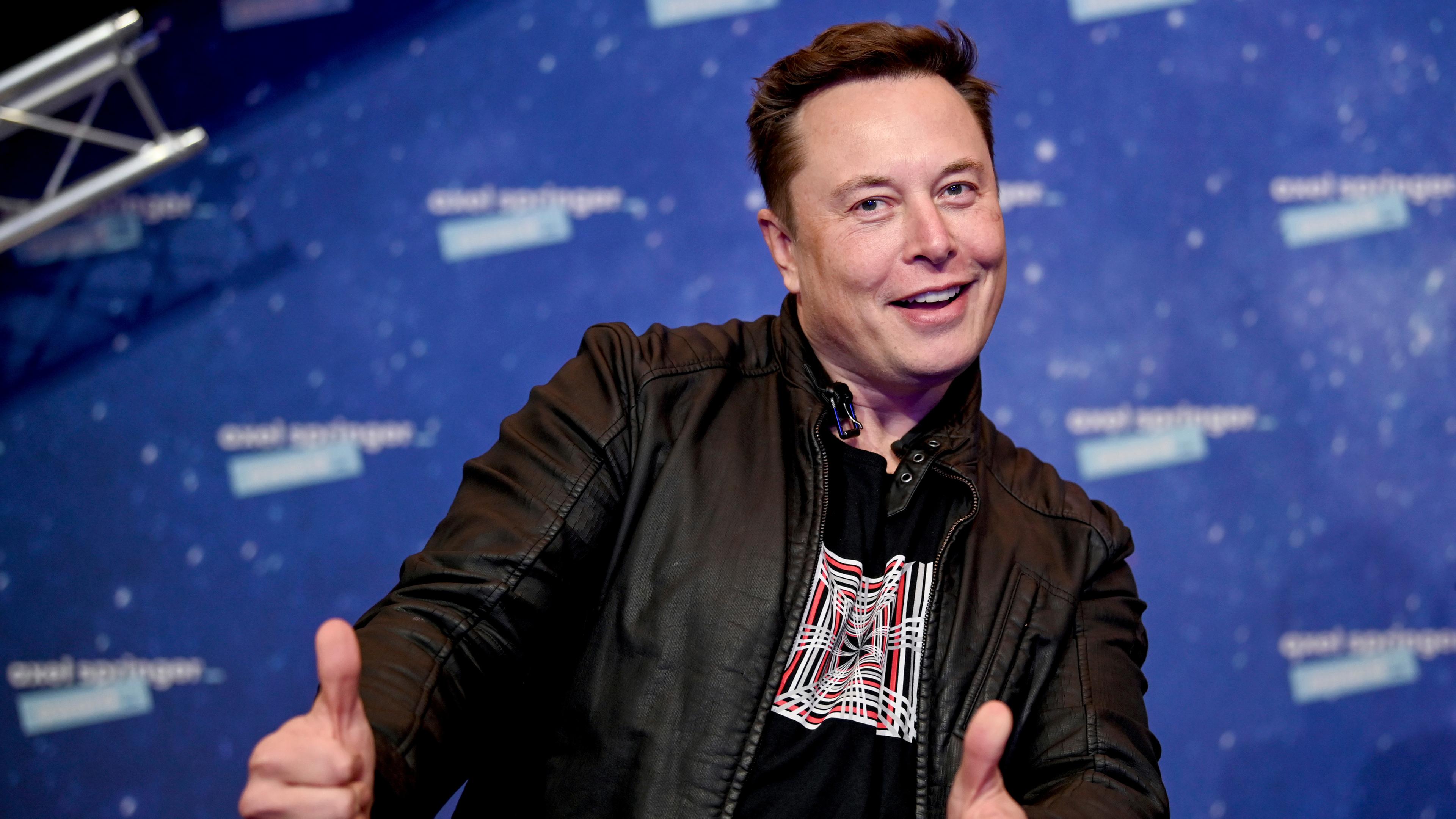 Elon Musk steht vor einer blauen Wand und zeigt zwei Daumen hoch in die Kamera.