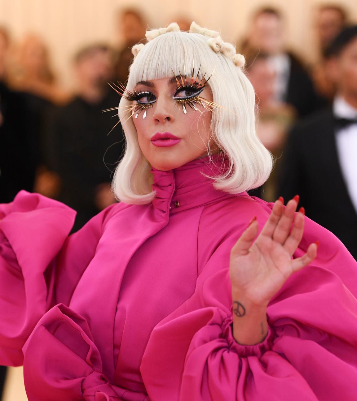  Lady Gaga trägt ein rosafarbenes Kleid mit Puffärmeln. Hellblonde Haare reichen ihr bis zur Schulter und sie trägt lange künstliche Wimpern. Im Hintergrund sind Fotografen und ein Mann im Smoking zu sehen.