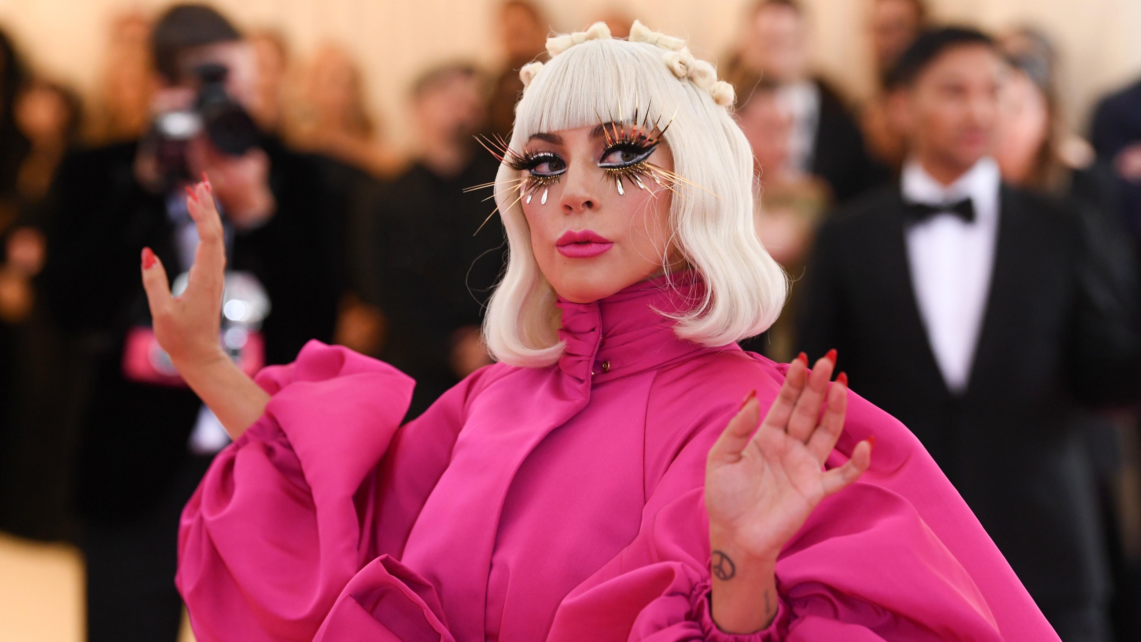  Lady Gaga trägt ein rosafarbenes Kleid mit Puffärmeln. Hellblonde Haare reichen ihr bis zur Schulter und sie trägt lange künstliche Wimpern. Im Hintergrund sind Fotografen und ein Mann im Smoking zu sehen.