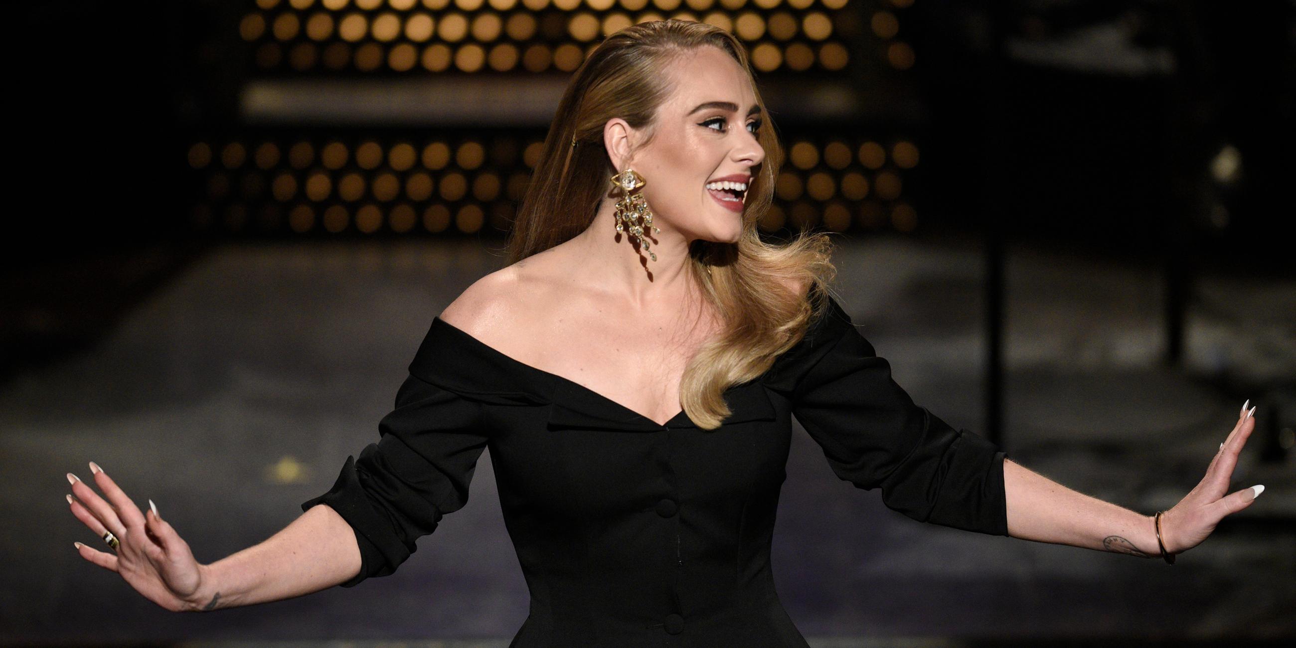Adele mit offenem Haar, einem schwarzen Kleid und großen Goldohrringen steht auf einer Bühne, schaut nach rechts und lächelt. Sie streckt die Arme aus.