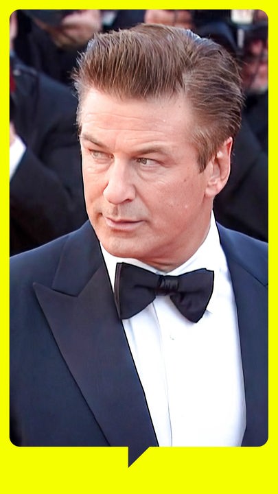 Alex Baldwin steht in Anzug und Fliege neben seiner dunkelhaarigen Frau. Beide schauen zur Seite, hinter ihnen stehen Fotografen.