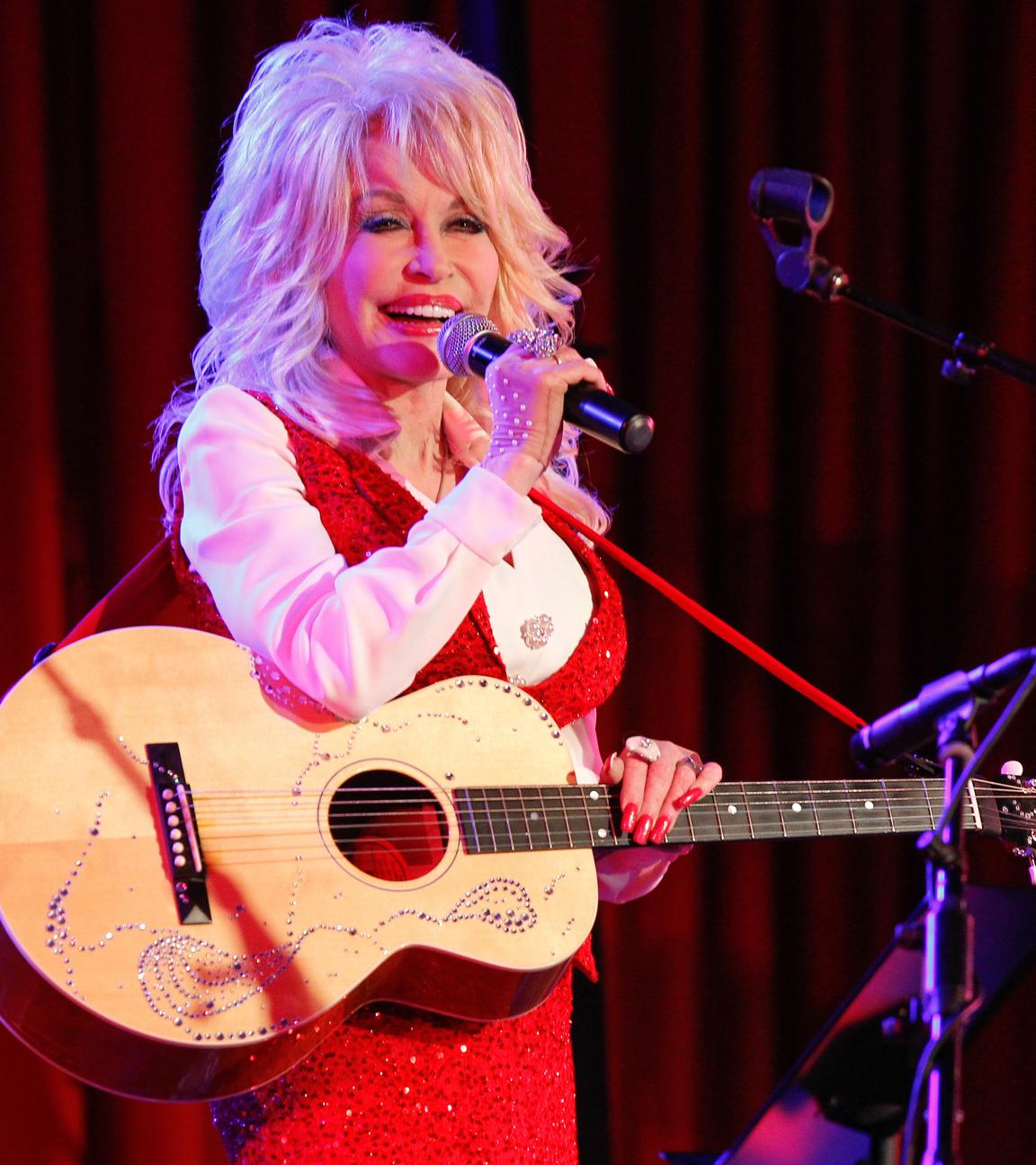 Dolly Parton hält in der linken Hand eine Gitarre. Sie steht vor einem roten Vorhang und singt in ein Mikrofon.